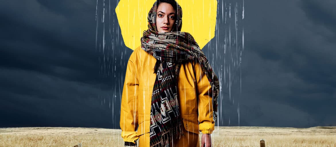 A woman stands with scarf around her head wearing a mustard jacket and carrying two suitcases. Behind her is a field of wheat with an old wooden fence. Behind her head is an illustration of a yellow sun with a cloud breaking with rain on top.