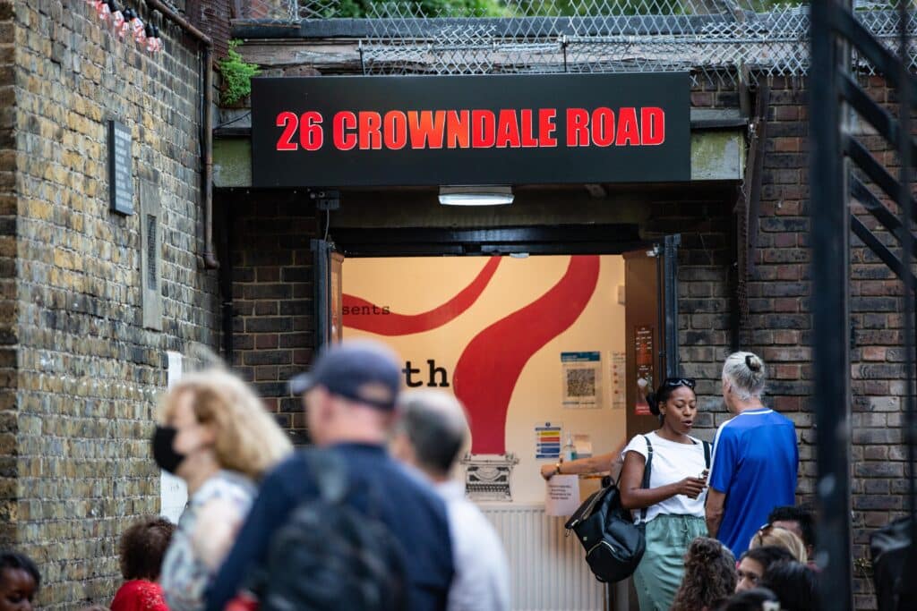 Theatre entrance to 26 Crowndale Road is visible with its doors open, people standing and chatting outside.