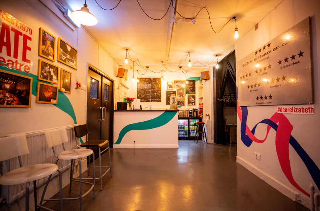 Foyer and bar are visible, with highchairs and decorative lights. The Gate Theatre’s branding, production photos and reviews are displayed on the walls, with swirls of colour surrounding. Photo taken during Dear Elizabeth run in summer 2021.