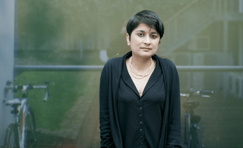 Full body shot of Shami Chakrabarti, stood in front of a bike rack with her hands in the pockets of her jeans. Also wearing a black blouse, black cardigan a string of pearls, hair cropped.