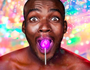 Close up of a smiling, wide-eyed, bare-chested black man, with a glowing purple lollipop in his mouth. The background behind him is bright, multi-coloured and full of sparkles